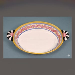 May include: An oval ceramic baking dish with a white interior and a yellow rim. The border features a pink wavy line pattern with blue accents. Two side handles are decorated with black and white stripes and a central pink circular accent.