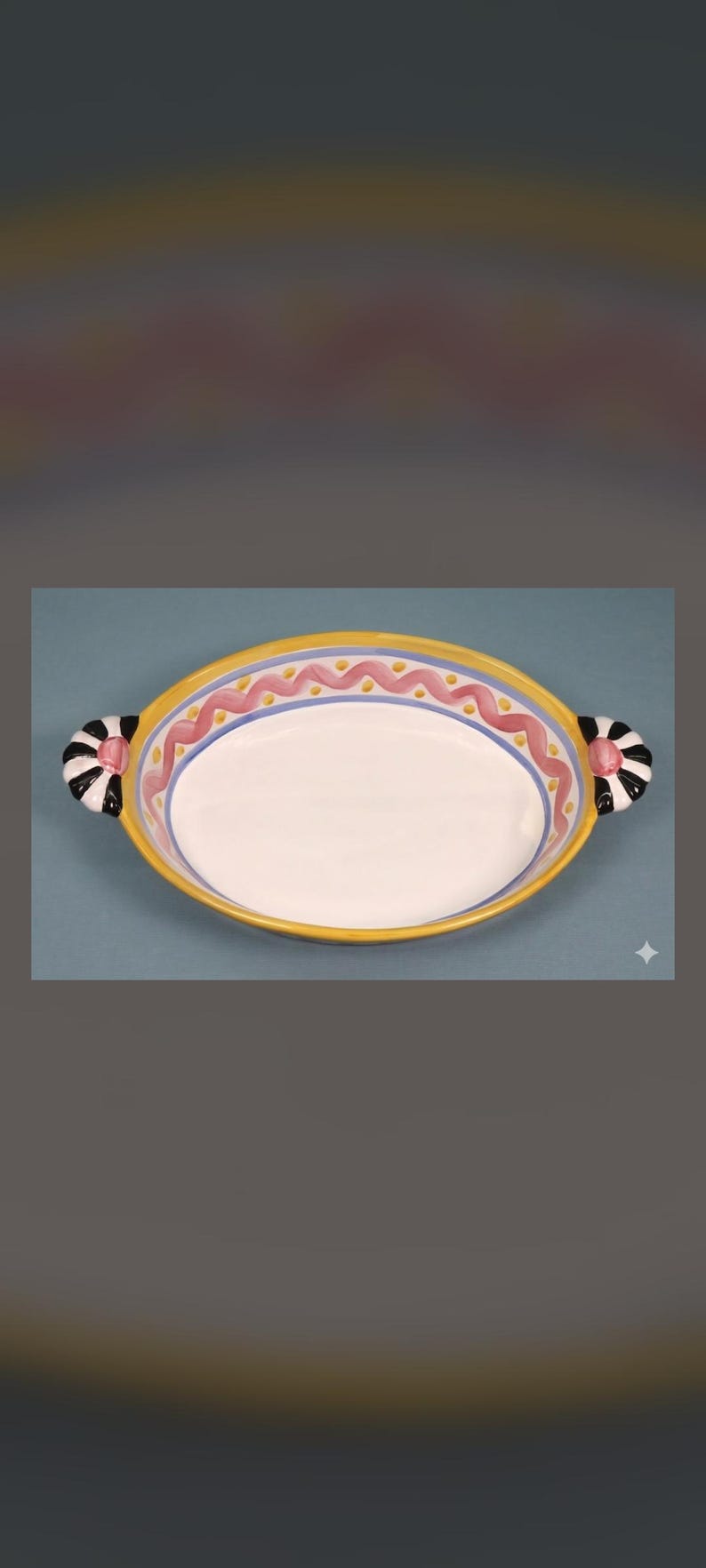 May include: An oval ceramic baking dish with a white interior and a yellow rim. The border features a pink wavy line pattern with blue accents. Two side handles are decorated with black and white stripes and a central pink circular accent.