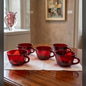 May include: A set of four translucent ruby red glass teacups with rounded bowls and curved handles. The cups are arranged in a cluster on a white fabric surface, showcasing a smooth, glossy finish and a consistent deep red color throughout.