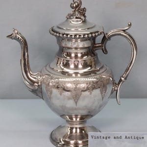 May include: A silver-colored antique teapot with a decorative handle and spout. The teapot has a lid with a detailed finial and is raised on a pedestal base. The body of the teapot features ornate engraved designs. The text "Vintage and Antique Estate Treasures" is visible.