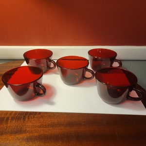 May include: Five ruby red glass cups with handles are arranged on a white surface. The cups have a simple, rounded design and are evenly spaced. The background is a solid red wall.