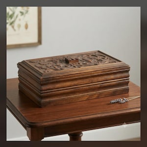 May include: A rectangular dark brown wooden box with a hinged lid featuring intricate carved floral and scrollwork patterns. The box has a tiered, stepped base design and a small square wooden knob centered on the top lid for opening.