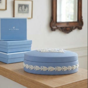 May include: A light blue round ceramic trinket box featuring white relief ornamentation. The lid displays a central white embossed design, while the side is decorated with a continuous white leaf garland. Three stacked light blue boxes with the Wedgwood logo are positioned behind the main item.