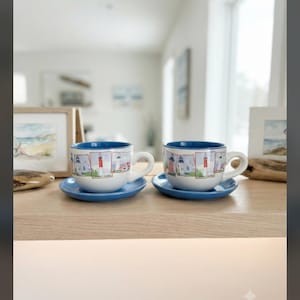 May include: Two white ceramic teacups with matching blue saucers, each featuring a series of colorful lighthouse illustrations around the exterior. The cups have a solid blue interior rim and a curved handle, resting on a light-toned wooden surface.