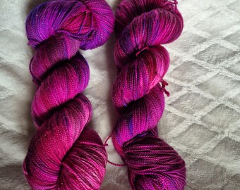 May include: Two skeins of vibrant, hand-dyed yarn in shades of pink, purple, and magenta. The yarn is twisted into a skein shape, with the text "Base Physalis" and "Base Samoa" in blue at the bottom. The yarn is on a white, textured surface.