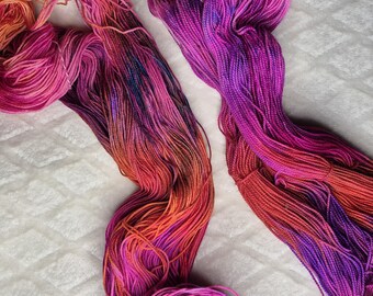 May include: Two skeins of hand-dyed yarn in vibrant shades of pink, orange, and purple. The yarn is arranged on a white, textured surface, showcasing the color variations and textures. Ideal for knitting or crochet projects.