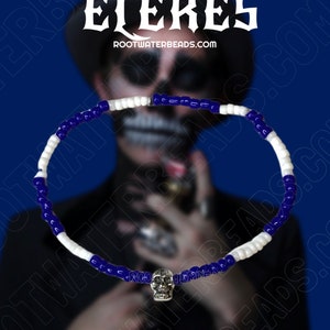 May include: A beaded necklace with alternating white and dark blue beads. The necklace has a silver skull pendant. The image includes the text "SHOP ELEKES" and "ROOTWATERBEADS.COM". The background is a dark blue.