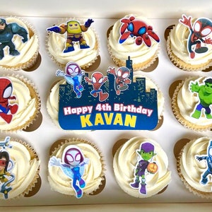 May include: A box of 10 cupcakes decorated with Spiderman and other superhero characters. The cupcakes are topped with white icing and have a variety of colourful decorations. The box has a sign that says "Happy 4th Birthday Kavan".