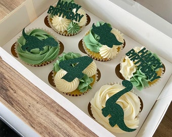 May include: A white cupcake box with six cupcakes decorated with green and white frosting. The cupcakes have dinosaur and "Happy Birthday" toppers.