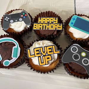 May include: Six cupcakes decorated with gamer-themed frosting and toppers. The cupcakes have brown wrappers and white frosting. The toppers include a blue and green headset, a black and white game controller, a blue and white Nintendo Switch, a black and white PlayStation controller, a yellow and black "Level Up" sign, and a yellow and black "Happy Birthday" sign.