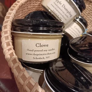 May include: A woven basket filled with several small glass jars of hand-poured soy candles. The labels read "Clove" and "Apple Dumpling". The jars have black lids and are from theprimitivebox.com, Schodack, NY.