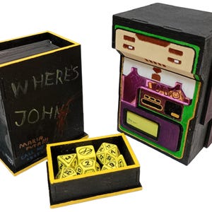 Pode incluir: Um conjunto de três acessórios de jogo de madeira. Inclui uma caixa preta com o texto "WHERE'S JOHN", uma caixa de dados com dados amarelos e uma caixa de jogo preta e verde com um design retrô. As caixas têm detalhes amarelos e roxos.