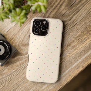 May include: A cream-coloured smartphone case featuring a pattern of small, multicoloured polka dots in shades of pink, blue, yellow, and green. The case has a rectangular cutout for a triple-lens camera system on the upper left corner.