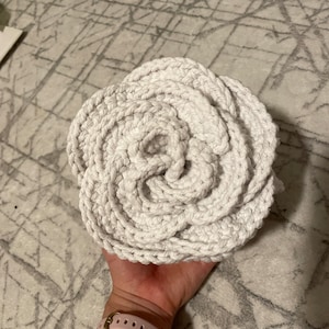以下が含まれることがあります： 厚手の糸で編まれた白いクロシェのバラが、手のひらに載せられています。バラは、中央に穴があり、複数のクロシェのループが重なったシンプルなデザインです。