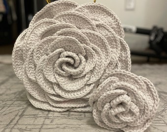 May include: A crocheted white rose-shaped handbag with a gold metal handle. A smaller white crocheted rose is sitting next to the bag.
