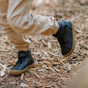 May include: Black leather ankle boots with brown soles. The boots have laces and a velcro strap. The boots are worn with tan pants. The boots are designed for children.