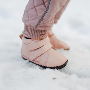 Könnte beinhalten: Nahaufnahme von einem Paar hellrosa Baby-Stiefel mit zwei Klettverschlüssen. Die Stiefel haben eine schwarze Sohle und werden mit einer passenden rosa Hose getragen. Die Stiefel stehen auf einer schneebedeckten Oberfläche.