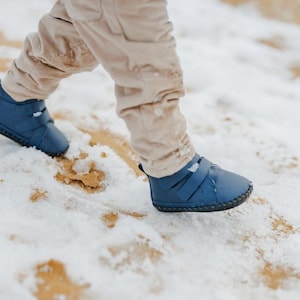 Könnte beinhalten: Marineblaue Kleinkinderschuhe mit Klettverschluss, getragen im Schnee. Die Schuhe haben eine runde Spitze und eine strukturierte Sohle. Das Kind trägt eine beige Hose.