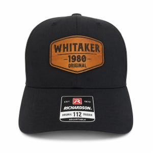 May include: A black baseball cap featuring a brown leather-like patch on the front with the text WHITAKER 1980 ORIGINAL. A rectangular sticker on the brim displays the text EST. 1970 RICHARDSON ORIGINAL 112 TRUCKER ADJUSTABLE with a red R logo.