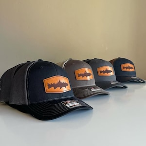 May include: Four trucker hats with a brown leather patch featuring a silhouette of a fish with trees on its back. The hats are in different colors: black, gray, brown, and navy blue.
