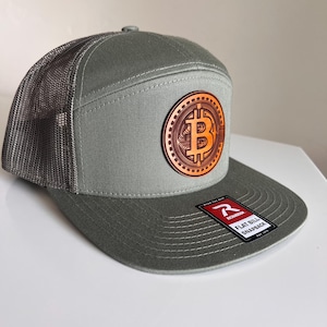 May include: A green and gray trucker hat with a brown leather patch featuring a Bitcoin symbol. The hat has a flat bill and a mesh back. The text "FLAT BILL SNAPBACK" is printed on a red tag attached to the hat.