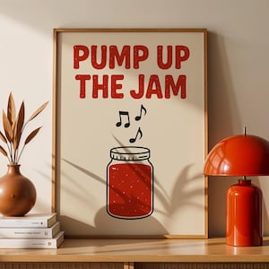 Puede incluir: Una impresión enmarcada con el texto "PUMP UP THE JAM" en letras rojas sobre un tarro de mermelada de dibujos animados con notas musicales. Una lámpara de mesa roja y un jarrón marrón con tallos decorativos están sobre un mueble de madera.