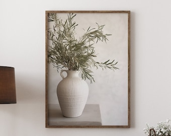 May include: A wooden dresser with six drawers and black handles. A framed print of a vase with greenery hangs above the dresser. A brown lamp with a tripod stand is on the left side of the dresser. A small potted plant is on the right side of the dresser. A woven basket with a book is on top of the dresser.
