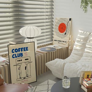 May include: A white couch with a white throw pillow. A wooden table with a record player and a black record on it. A black framed poster with the text 'COFFEE CLUB' and a coffee maker illustration. A white framed poster with the text 'HERE COMES THE SUN' and a red circle with a black line through it. A white lamp with a white shade. A brown leather chair with a white rug underneath.