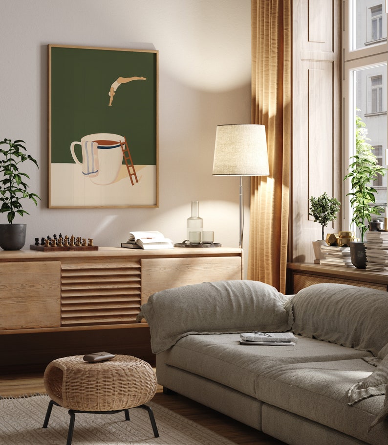May include: A living room interior with a light gray sofa, a wooden cabinet, a wicker ottoman, and a floor lamp with a beige lampshade. A framed print of a stylized illustration of a person diving into a coffee mug hangs on the wall.