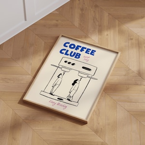 May include: A framed poster with a white background and black illustrations. The poster says 'COFFEE CLUB' in blue text with the words 'served daily' in smaller blue text. The poster shows two people drinking coffee from a coffee machine. The poster says 'Every Morning' in pink text.