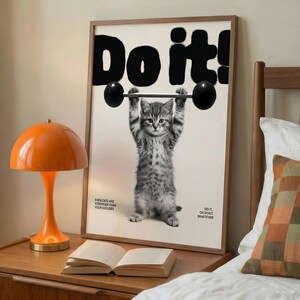May include: A framed print featuring a kitten lifting a barbell. The words "Do it!" are above the kitten, with the phrases "Even cats are stronger than your excuses" and "Do it... or don't whatever" below. The print is on a nightstand with an orange lamp and an open book.