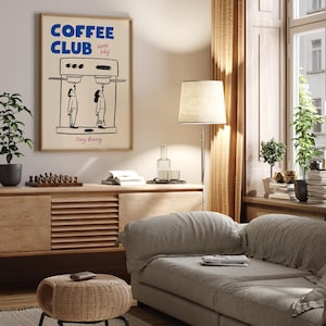 May include: A living room interior with a light brown couch, a wooden side table, a wicker stool, a floor lamp with a white lampshade, and a framed poster with the text 'COFFEE CLUB Served Daily Every Morning' and a cartoon illustration of a coffee machine with two people drinking coffee.