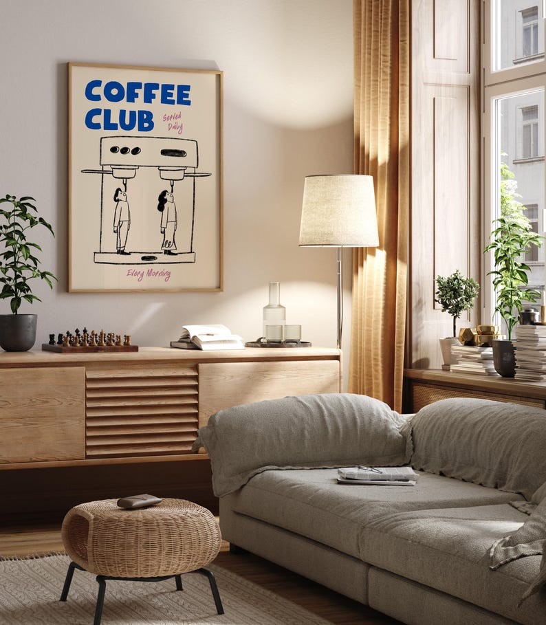 May include: A living room interior with a light brown couch, a wooden side table, a wicker stool, a floor lamp with a white lampshade, and a framed poster with the text 'COFFEE CLUB Served Daily Every Morning' and a cartoon illustration of a coffee machine with two people drinking coffee.