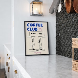 May include: A black framed poster with white text that reads 'COFFEE CLUB Served Daily Every Morning' and a black and white illustration of a coffee maker with two people standing underneath it.