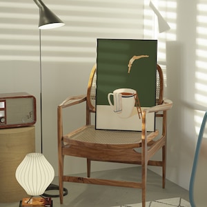 May include: A wooden chair with a woven cane seat and back, a green framed print of a person diving into a cup of coffee, and a white lamp with a white shade.