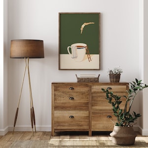 May include: A framed illustration of a person diving into a large coffee mug with a ladder. The illustration is on a green background and the mug is white with a blue and white towel around it. The frame is brown wood.