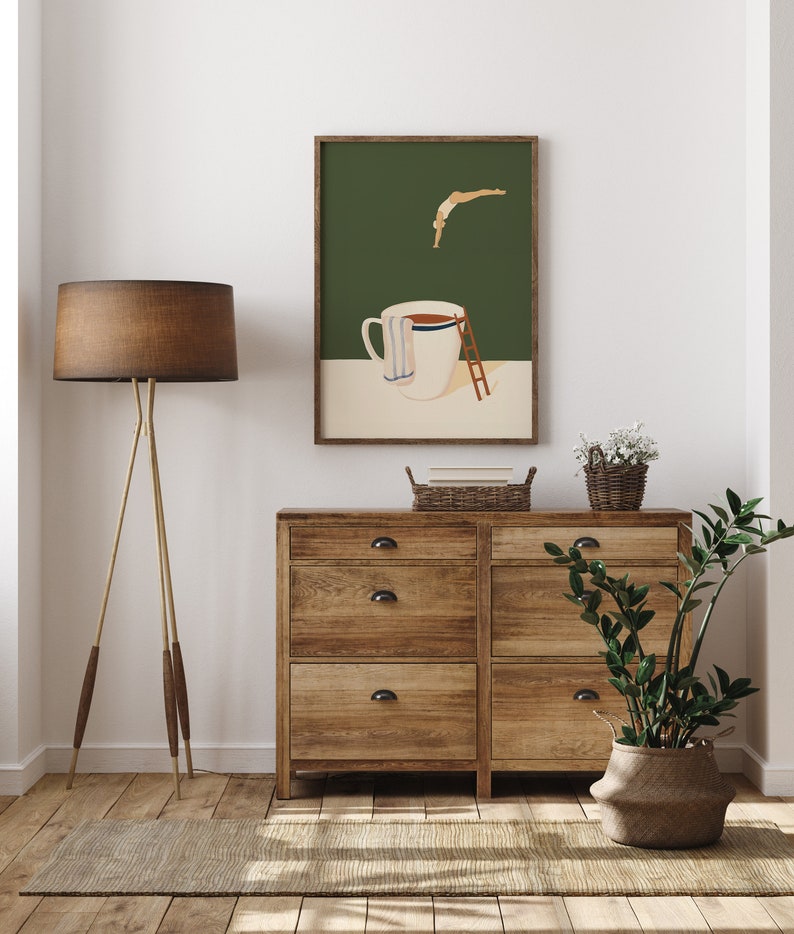 May include: A framed illustration of a person diving into a large coffee mug with a ladder. The illustration is on a green background and the mug is white with a blue and white towel around it. The frame is brown wood.