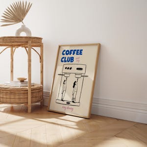 May include: A framed print with a light beige background. The print features a black and white illustration of a coffee maker with two people standing underneath it. The print has the text 'COFFEE CLUB' in blue letters and 'Special Daily' in smaller black letters. The print also has the text 'Every Morning' in pink letters.