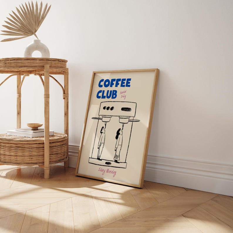 May include: A framed print with a light beige background. The print features a black and white illustration of a coffee maker with two people standing underneath it. The print has the text 'COFFEE CLUB' in blue letters and 'Special Daily' in smaller black letters. The print also has the text 'Every Morning' in pink letters.