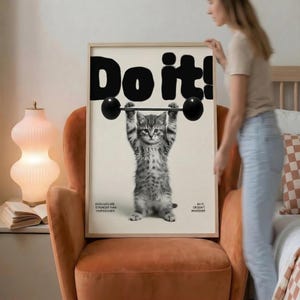 May include: A framed print featuring a gray tabby kitten lifting a barbell. Bold black text above reads "Do it!". Additional text states "Even cats are stronger than your excuses" and "Do it or don't whatever". The print is in a light-colored wooden frame.