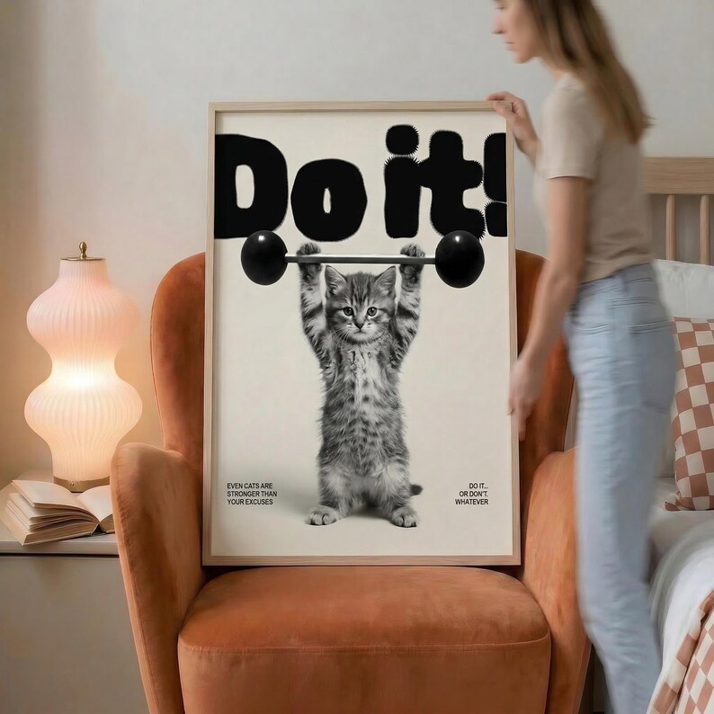 May include: A framed print featuring a gray tabby kitten lifting a barbell. Bold black text above reads "Do it!". Additional text states "Even cats are stronger than your excuses" and "Do it or don't whatever". The print is in a light-colored wooden frame.