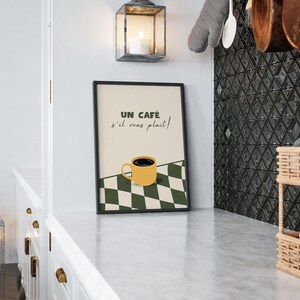 May include: A black framed print with a white and green chequered background. The print features a yellow coffee cup with black coffee and the text "Un caf&eacute; s'il vous pla&icirc;t!" in French.