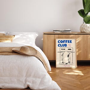 May include: A poster with a white background and black line drawings of two people standing in front of a coffee maker. The poster has the text 'COFFEE CLUB' in blue letters and the text 'Every Morning' in black letters.