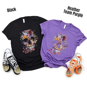 Pu&ograve; includere: Due magliette, una nera e una viola, con un design a teschio floreale. La maglietta nera ha scarpe da ginnastica bianche e nere, e quella viola ha scarpe da ginnastica arancioni. Il testo "Black" e "Heather Team Purple" &egrave; visibile.