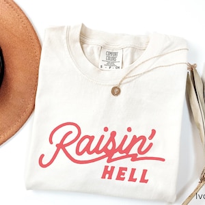 May include: A white t-shirt with the text "Raisin' Hell" in red lettering. The shirt is laying flat on a white surface with a brown hat and a dried palm leaf in the background.