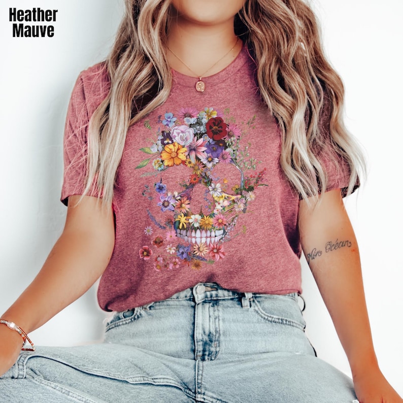 Pu&ograve; includere: T-shirt color malva melange con un design a teschio floreale. Il teschio &egrave; composto da fiori colorati nei toni del rosa, giallo, viola e rosso. Il testo "Heather Mauve" &egrave; nell'angolo in alto a sinistra.