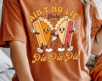 May include: Rust-colored t-shirt with a retro graphic of two pie slices with faces, arms, and legs. The text reads "Ain't No Lie Baby" above and "Pie Pie Pie" below. The pie slices are giving peace signs.