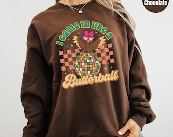 May include: A dark chocolate brown long-sleeve sweatshirt featuring a graphic print of a turkey wearing pink sunglasses and a bow, standing on a colorful disco ball. The text reads I came in like a Butterball with a checkered background pattern.