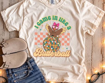 May include: An ivory short-sleeve t-shirt featuring a graphic of a brown turkey wearing pink heart-shaped sunglasses and a pink bow, perched on a colorful disco ball. The text reads I came in like a Butterball in a retro font style.