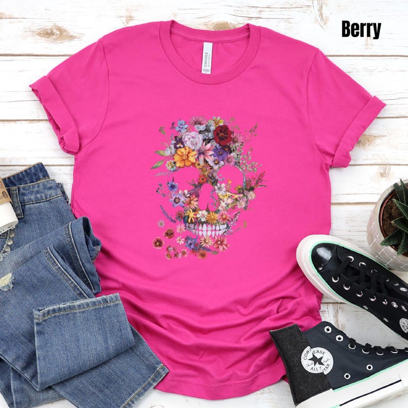 Pu&ograve; includere: Una maglietta rosa acceso con un design a teschio floreale. Il teschio &egrave; composto da vari fiori colorati. La maglietta &egrave; abbinata a jeans blu e scarpe da ginnastica nere. La parola "Berry" &egrave; visibile nell'angolo in alto a destra.